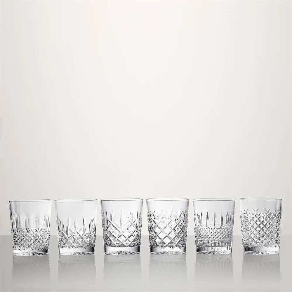 Six clear cut-crystal tumblers with different diamond and geometric patterns, lined up in a row on a reflective white surface against a light gray background from our Waterford Crystal Australia – Outlet & Sale Collection