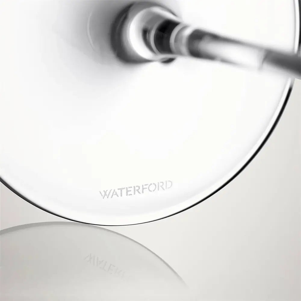 Close-up of a Waterford table lamp base showing a circular white shade, a slim metallic stem, and the Waterford logo on a reflective light gray surface.